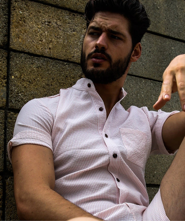 Man wearing a light pink shirt with black buttons against a stone wall.